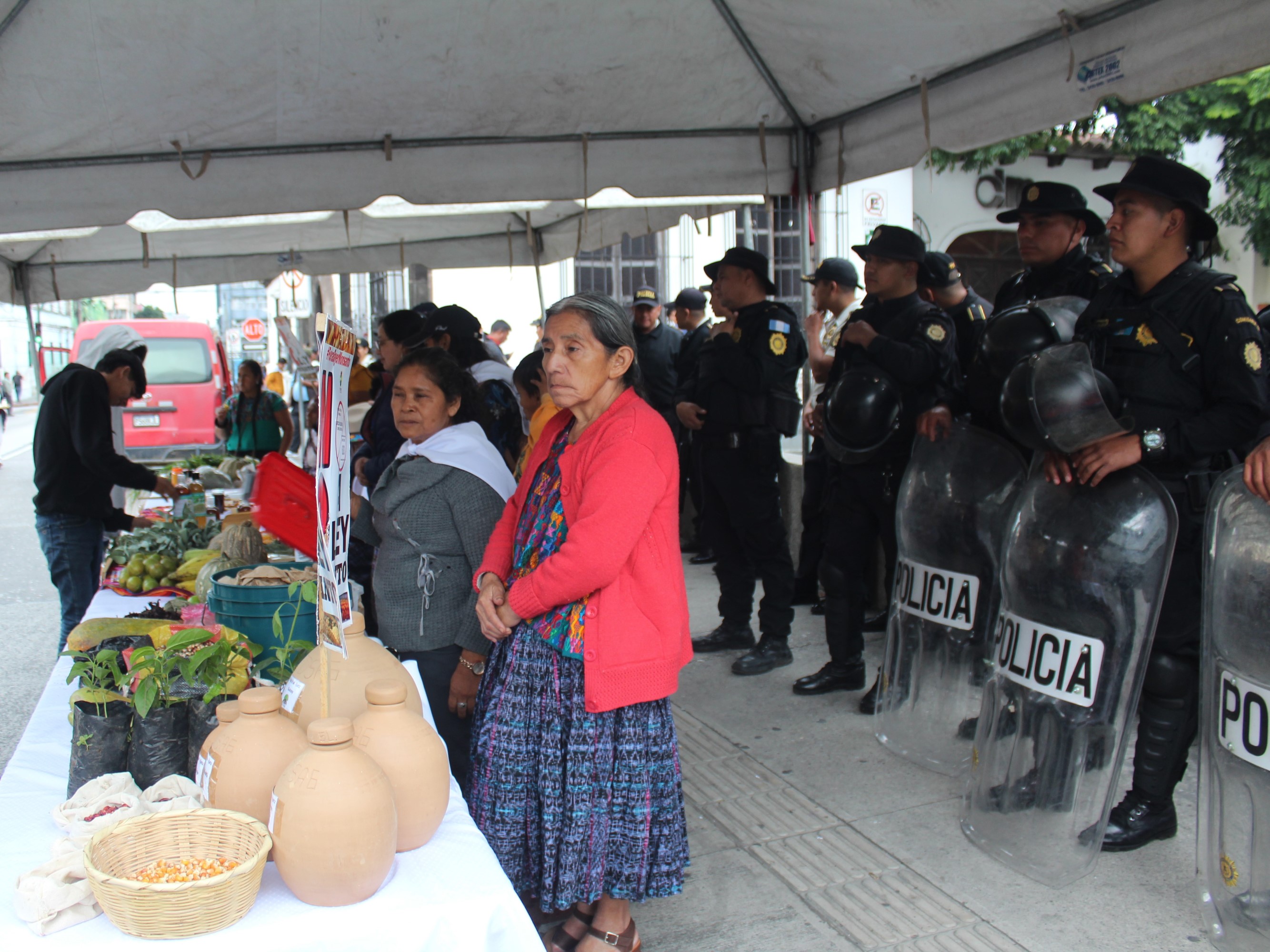 Women stand at a table of jars and foods at an outdoor market with uniformed policemen in full riot gear stand five feet behind them.