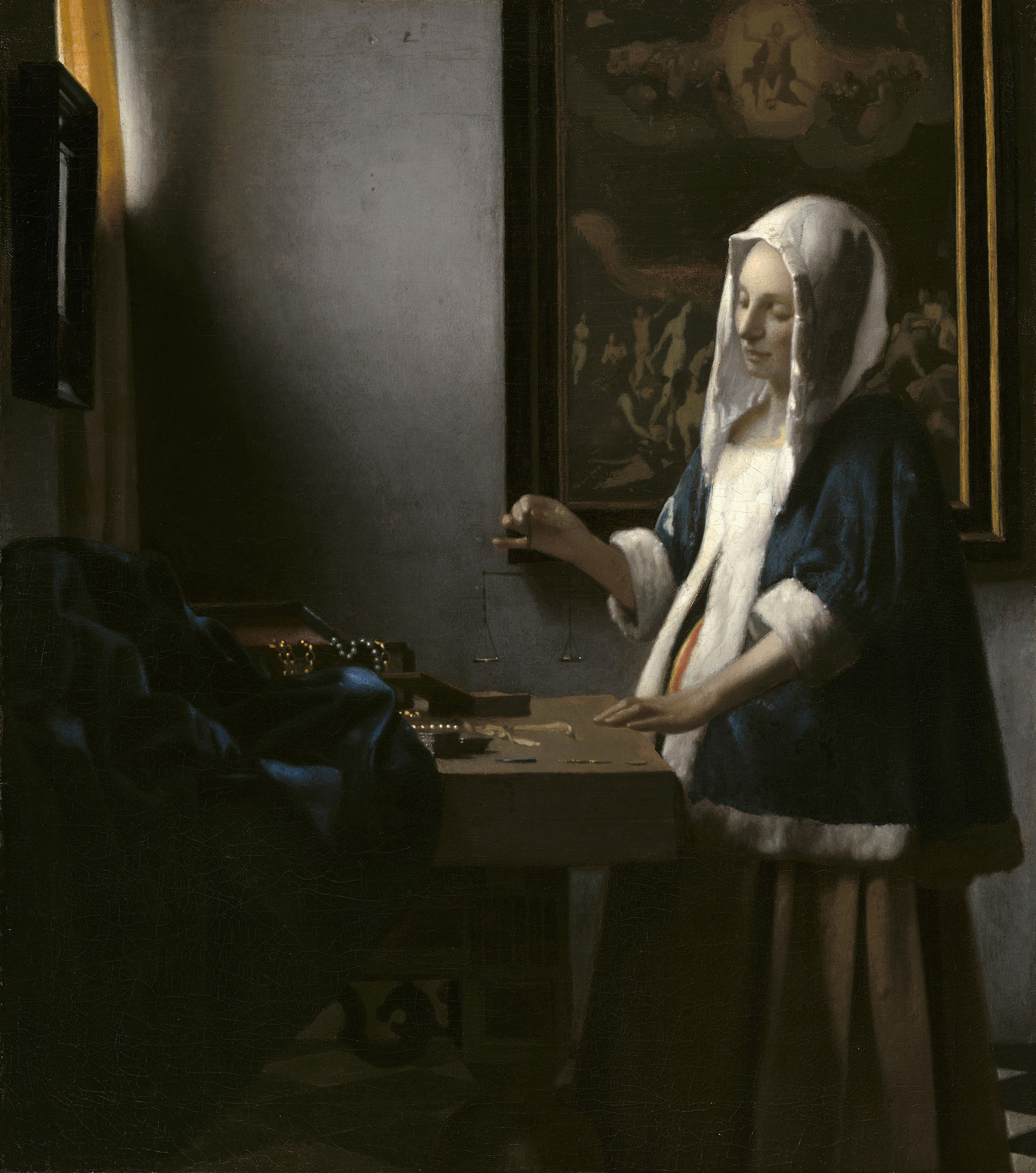A young woman with pale skin, wearing a velvety blue robe trimmed with white fur, stands before a small table and an open jewely box that holds a few strands of pearls. She holds a delicate set of scales in her hand, and is illuminated by angled light from a high window.