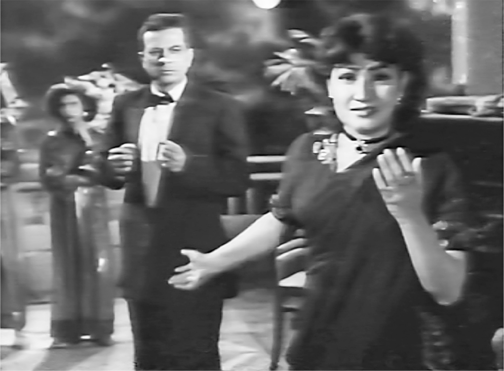 A medium interior close-up of a woman dressed in an elegant black sari facing and gesturing towards the camera. Behind her on the left is a man dressed in a tuxedo, also facing the camera.