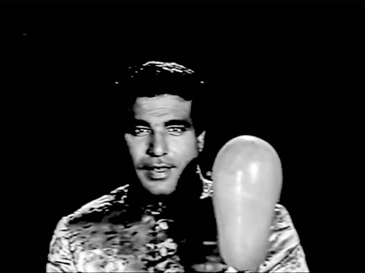 A dark background frames a close-up shot of a young man wearing a fine wedding sherwani and facing the camera, with a vertically elongated balloon floating upwards in front of his left shoulder.