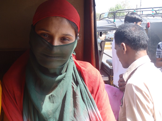 A photograph of a woman sitting in the back of an autorickshaw, with an orange scarf covering her hair and shoulders and a thin green scarf covering most of her face except for her eyes. Other people and vehicles can be seen in the background.