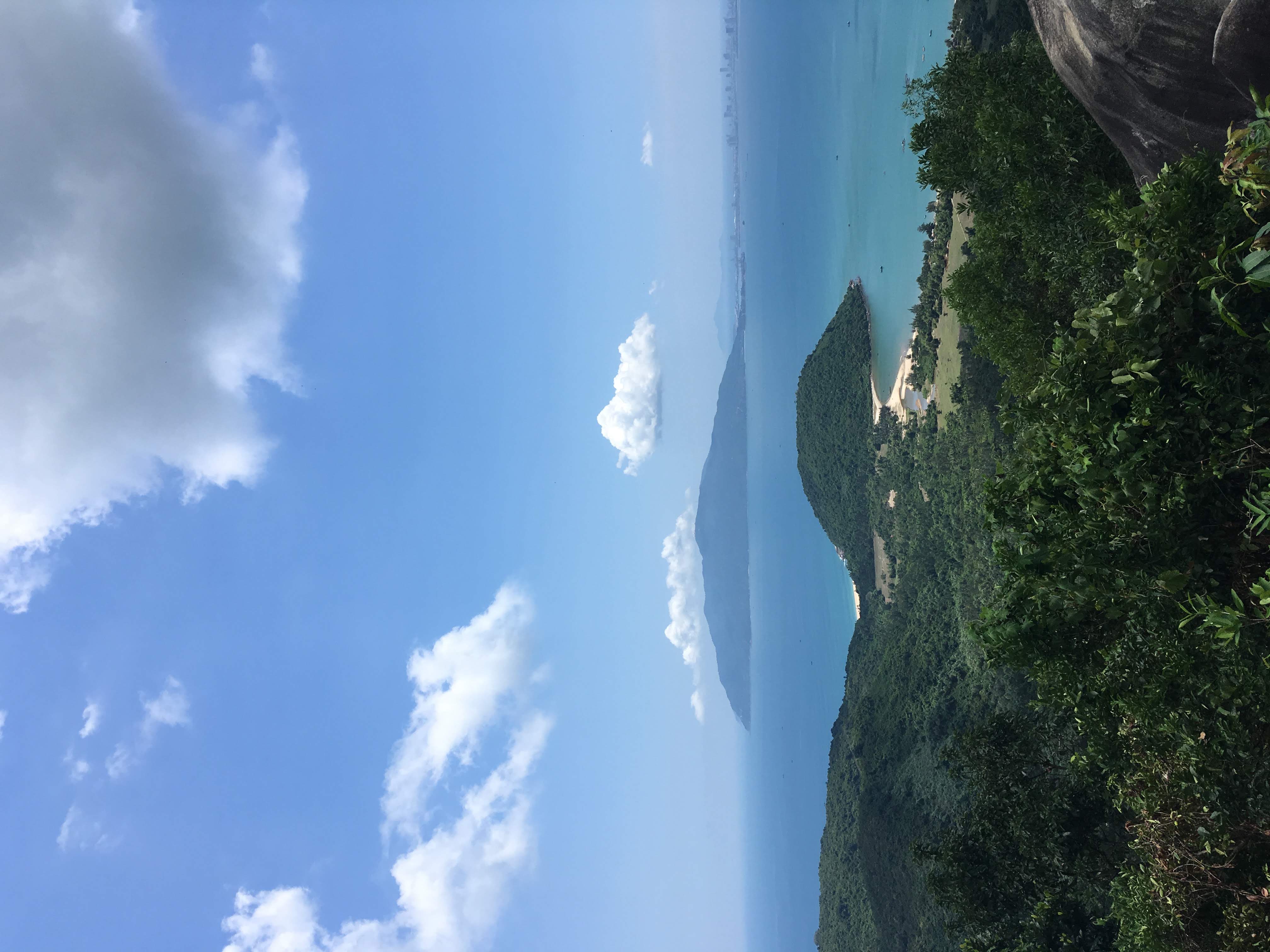A view over green hills to a blue sea, with an island in the distance, under a blue sky dotted with clouds.