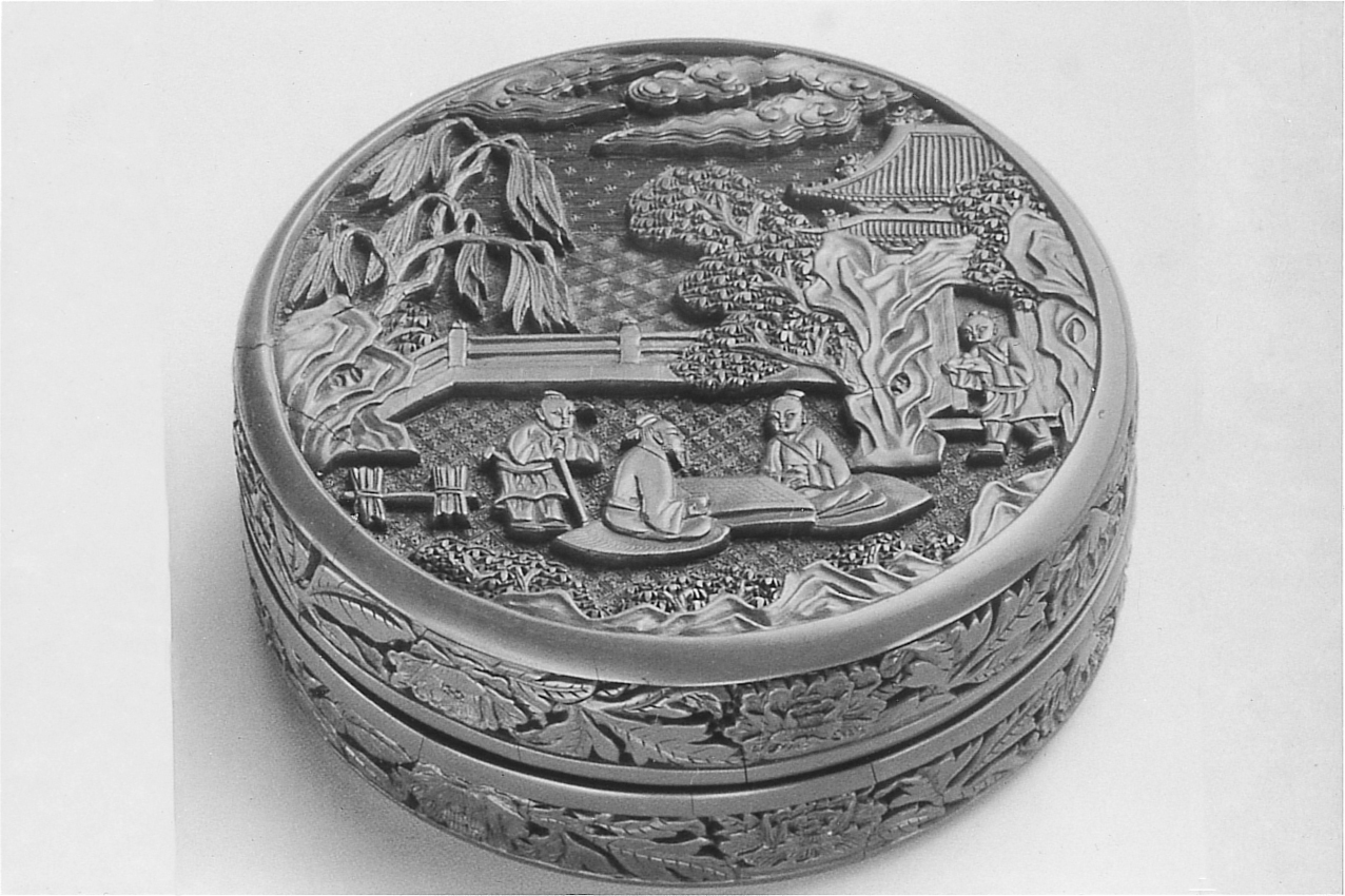 A photograph of a round, intricately carved, covered box, with a scene of three scholars talking in a garden on the lid, and foliage around the edges.