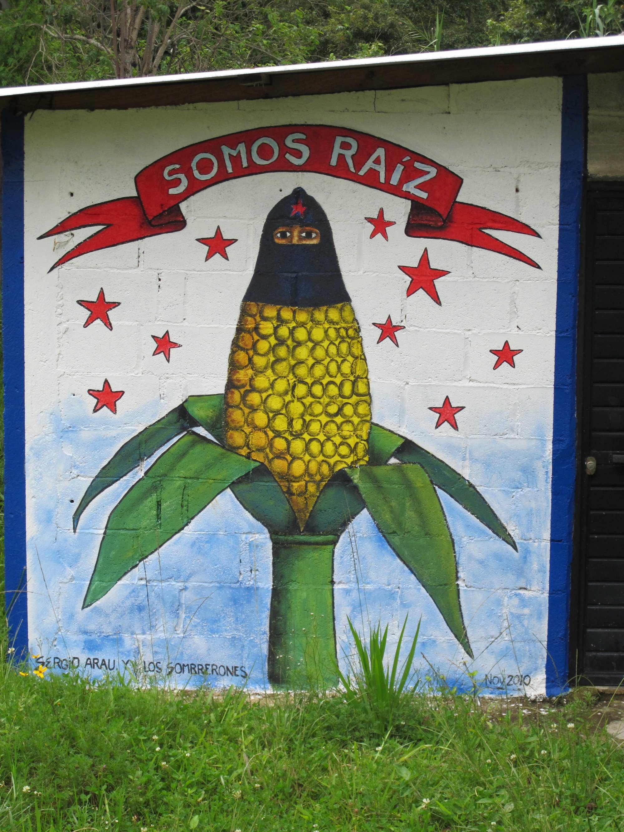 Painted mural of a maize cob with downturned husks, kernels, and hooded face with eyes showing.