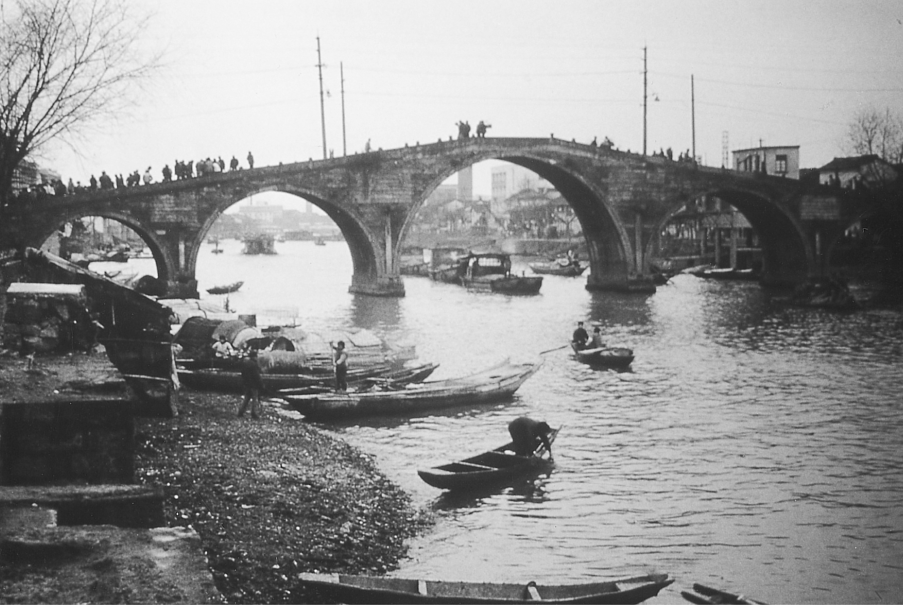 A photograph of a bridge built of five stone arches over a canal, with a high central arch and smaller ones on each side. A few small boats navigate the canal, while others line the shores. People cross the bridge on foot.