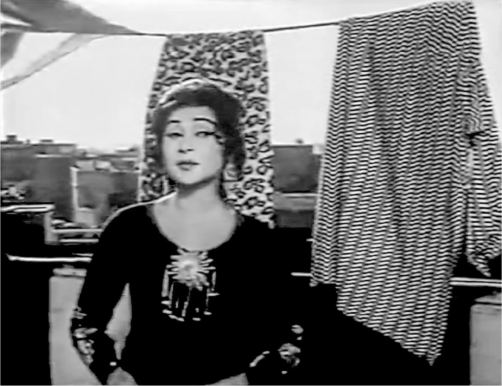 A medium close-up of a young woman in an embroidered black dress standing outside on a rooftop, facing the camera. Clothes hang on a clothesline behind her and in the distance beyond that is the dense rooftop cityscape of the Walled City in Lahore.