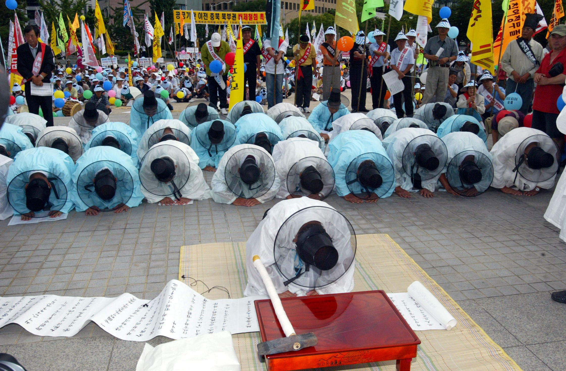 This is a contemporary photograph showing three rows of elderly Korean men wearing the traditional blue or white robes and black hats of Confucian scholars, prostrate, with heads bowed. In front of this group, one of the men is on his own. He kneels on a mat, behind a small carved and lacquered orange wooden table, with a wood-handled ax set upon it. A long scroll between the man and the table unrolls to the left beyond the edge of the photograph. Behind these men are a group of standing and sitting protesters in street clothes. Some protesters are holding flags on poles, some are holding picket signs, and some have sashes across their chests. Many wear blue gowns and white hats.