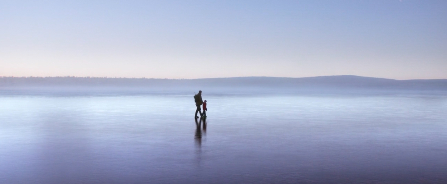 Long-distance view of an adult and a child walking across the surface of a frozen lake, with forested hills in the distant background. 