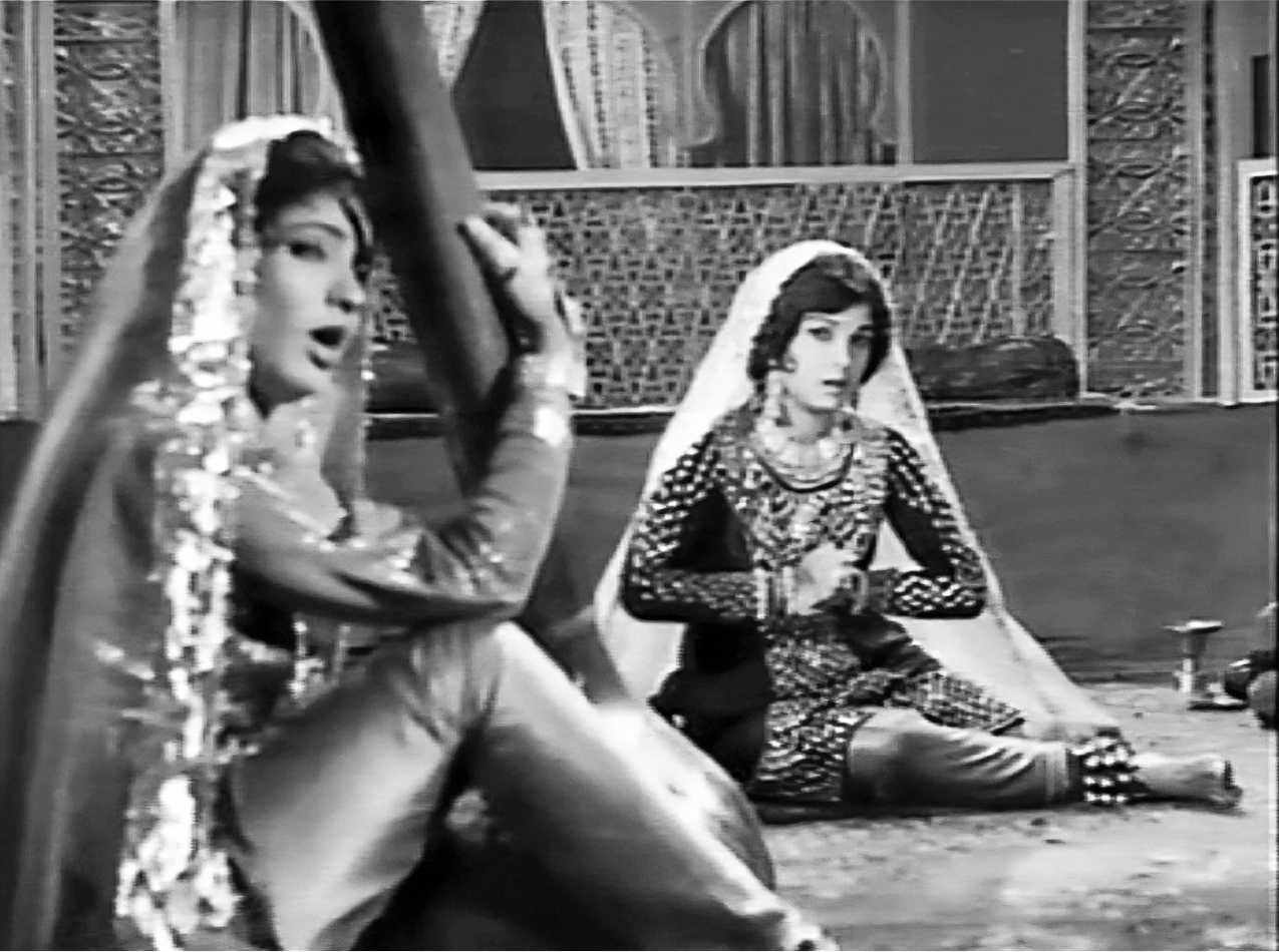 A traditionally decorated interior space with two seated women dressed in North Indian dancing outfits. The woman on the left holds a large string instrument, while the woman on the right sits in a dance pose with ankle-bells on her feet.