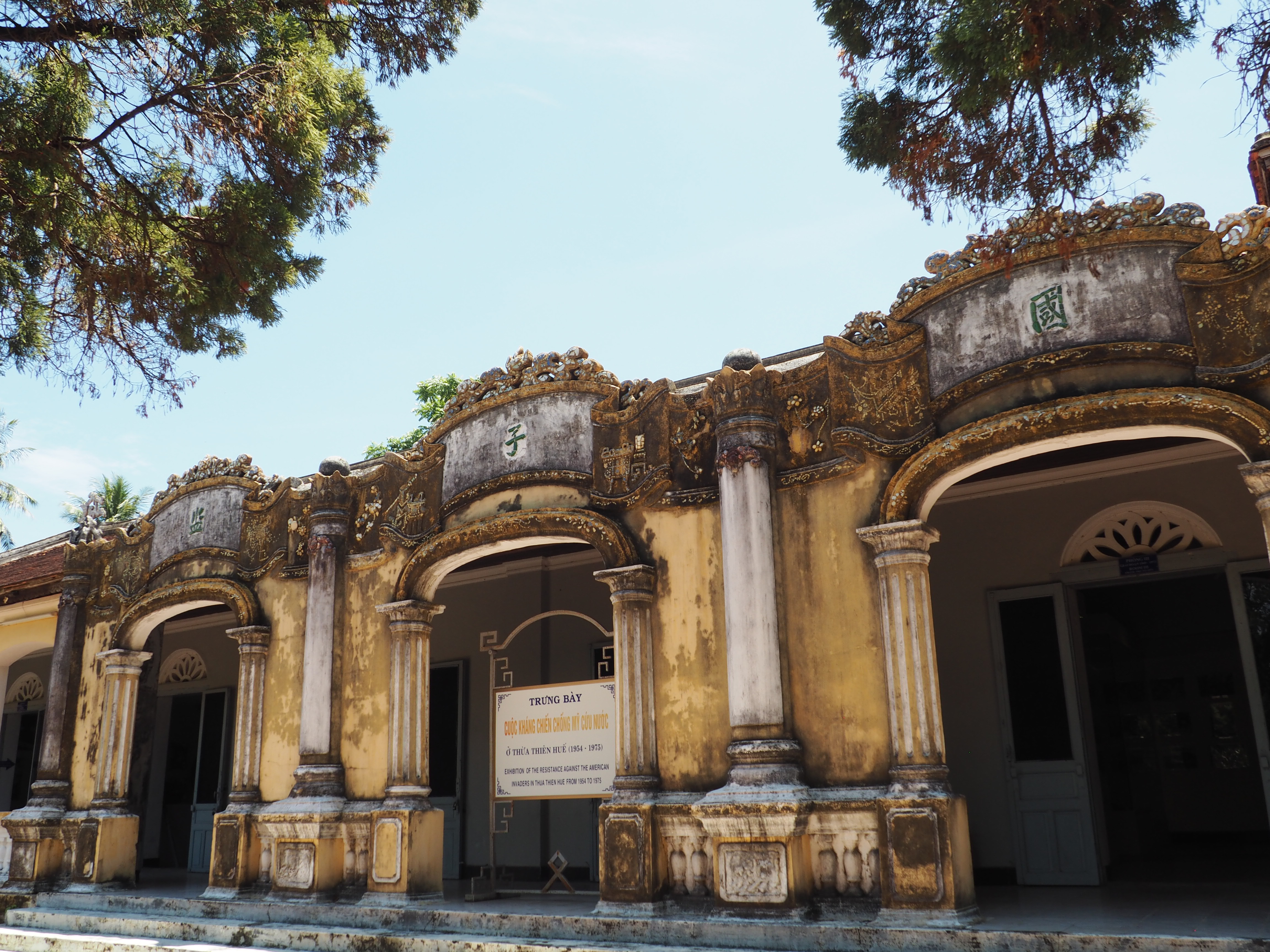 A building with a weathered yellow façade composed of a row of arches separated by columns. A Chinese character is inscribed in a space above each arch.
