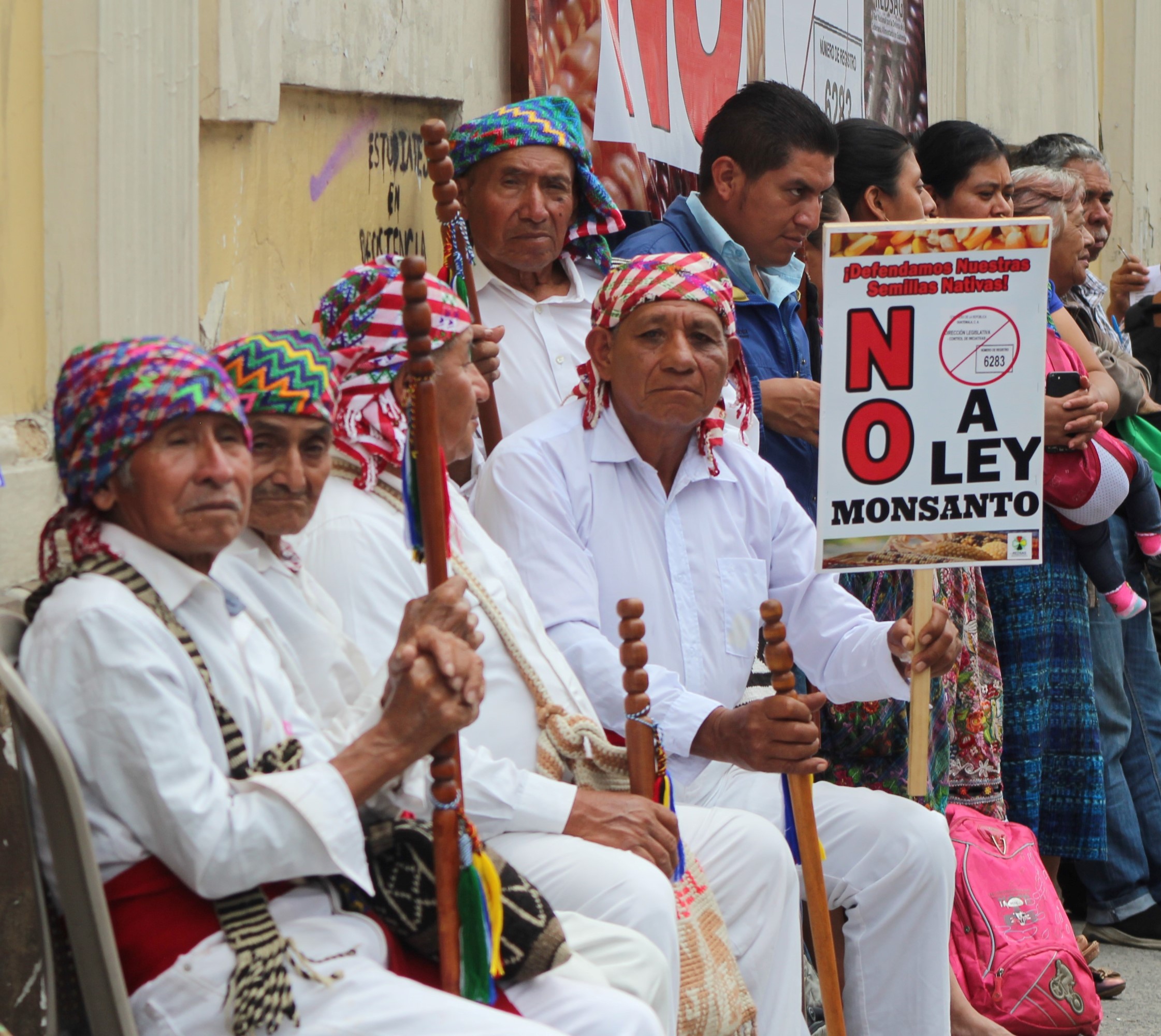 Five men holding staffs and dressed in white shirts and pants with woven scarves wrapped around their heads sit against a building. One of them holds a sign with a large “No.”