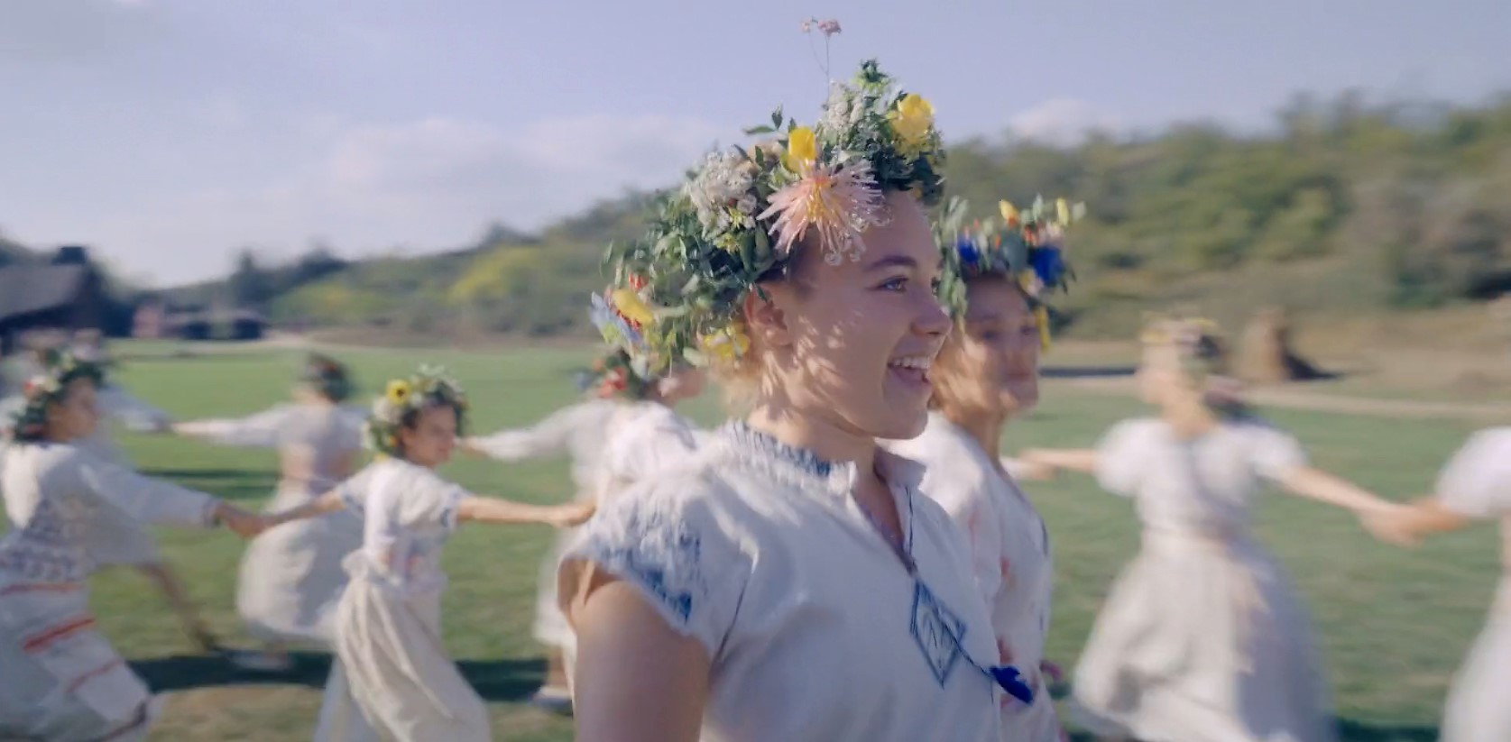A close-up view of a young woman smiling with flowers in her hair and dressed in a white, rustic dress as she joins hands with a group of similarly dressed women in a traditional folk dance.