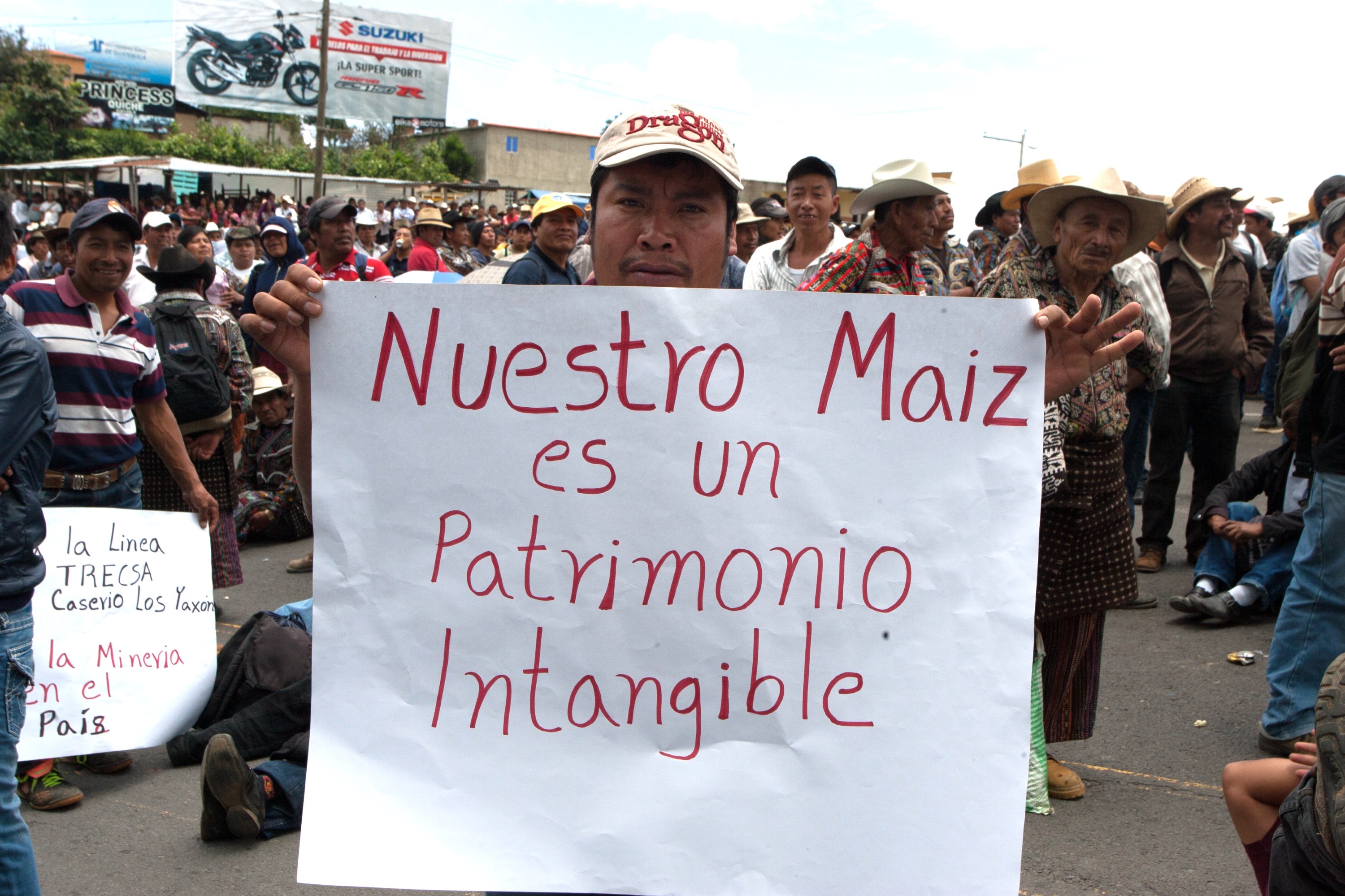 A close up of man in a baseball cap holding a homemade sign that says, “Our maize is intangible patrimony.” He is standing at the edge of a crowd of people.