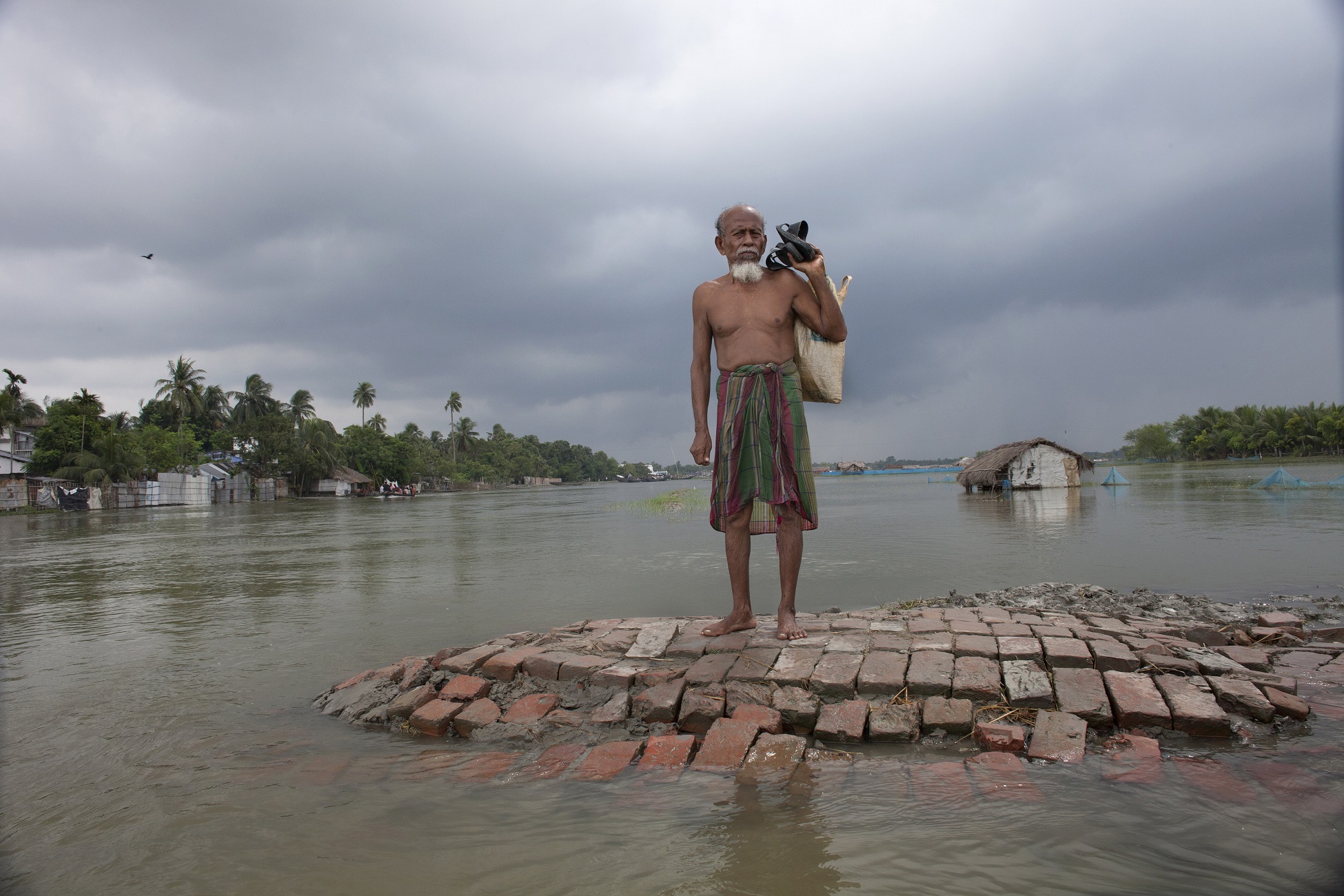 An older man with a white beard and bare chest stands barefoot, with his sandals in his hands, on a small bit of land in the middle of an overflowing river, under dark clouds. Half-flooded buildings are visible in the background.