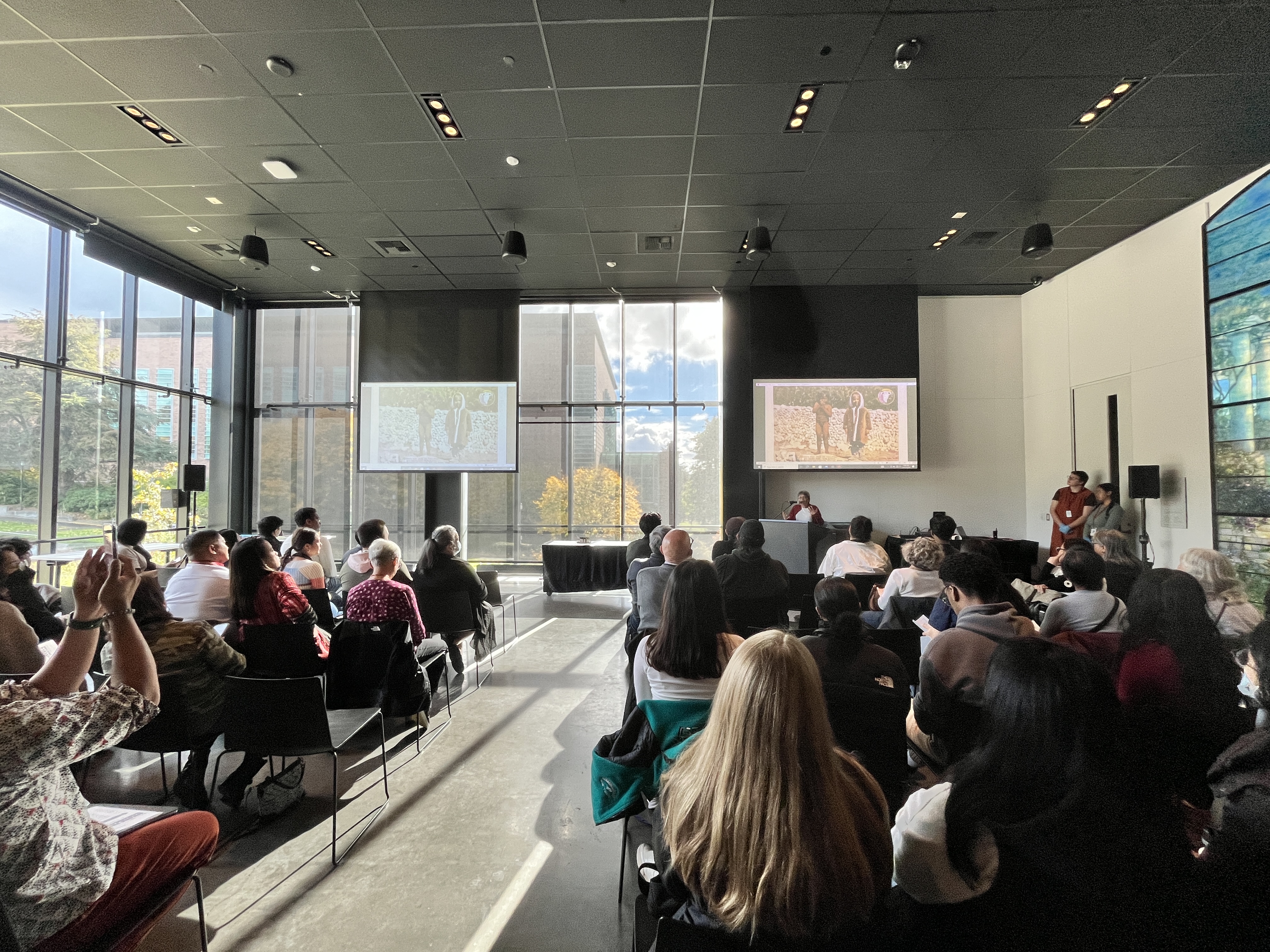 Building the Barangay event in a glass room, with one person presenting to a seated crowd in front of two projector screens.