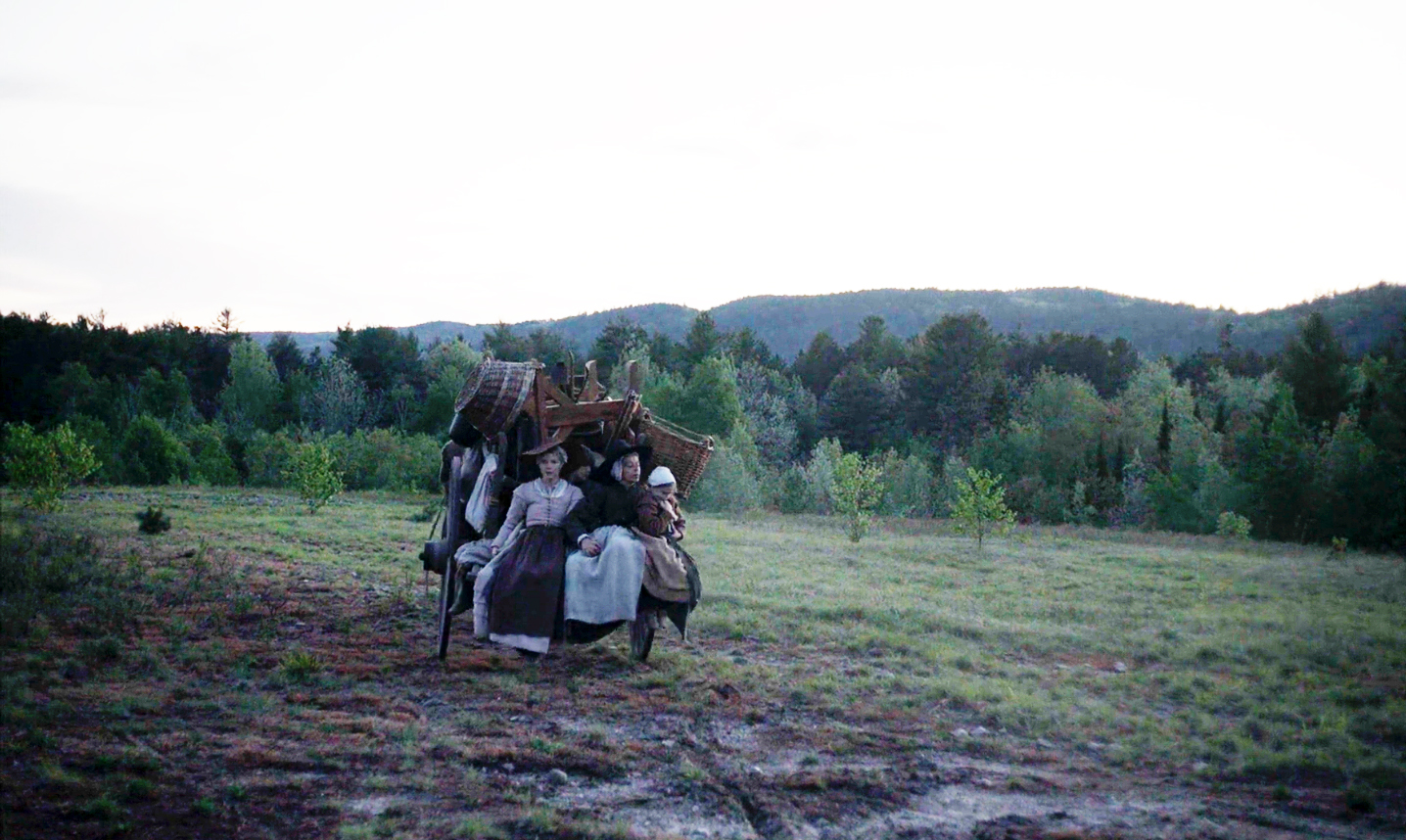 A family of Puritan settlers in colonial America ride on the back of a horse-drawn carriage that is heading into a densely forested wilderness landscape in the background.