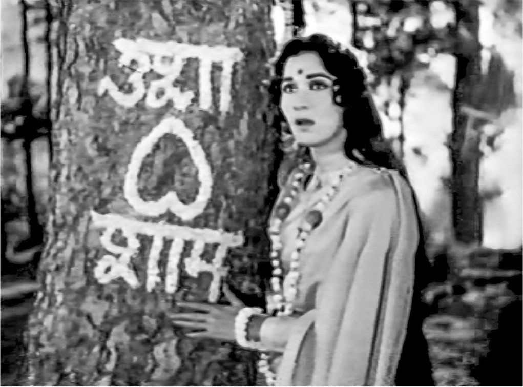 An exterior scene of a woman dressed in a white sari with a forehead bindi, long hair, and a garland of flowers around her neck. She stands next to a tree trunk, facing the camera, and points towards a large white inscription in Hindi on the trunk, and which has an upside-down symbol of a heart between two lines of text.