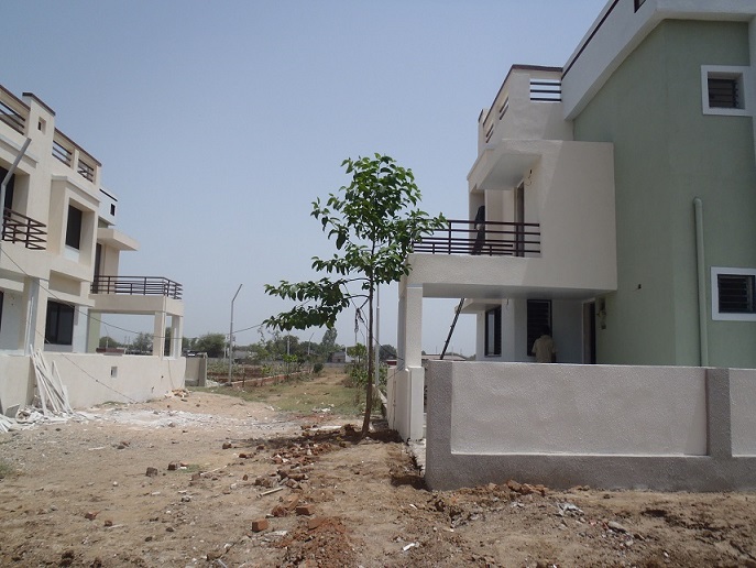 Two new two-story houses, built in a modernist style and painted white, sit on a dirt road. A small tree grows beside one of the houses, but the ground is otherwise bare.