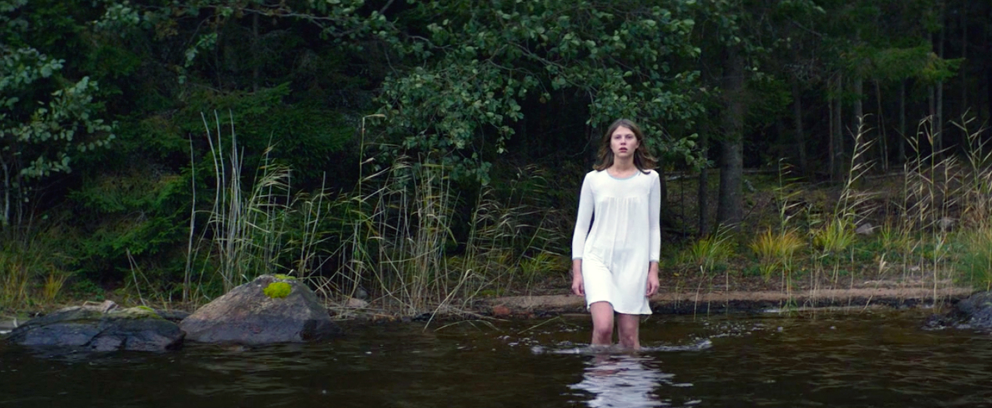 A young woman dressed in a white nightgown walks into a lake with a serious and determined look on her face.