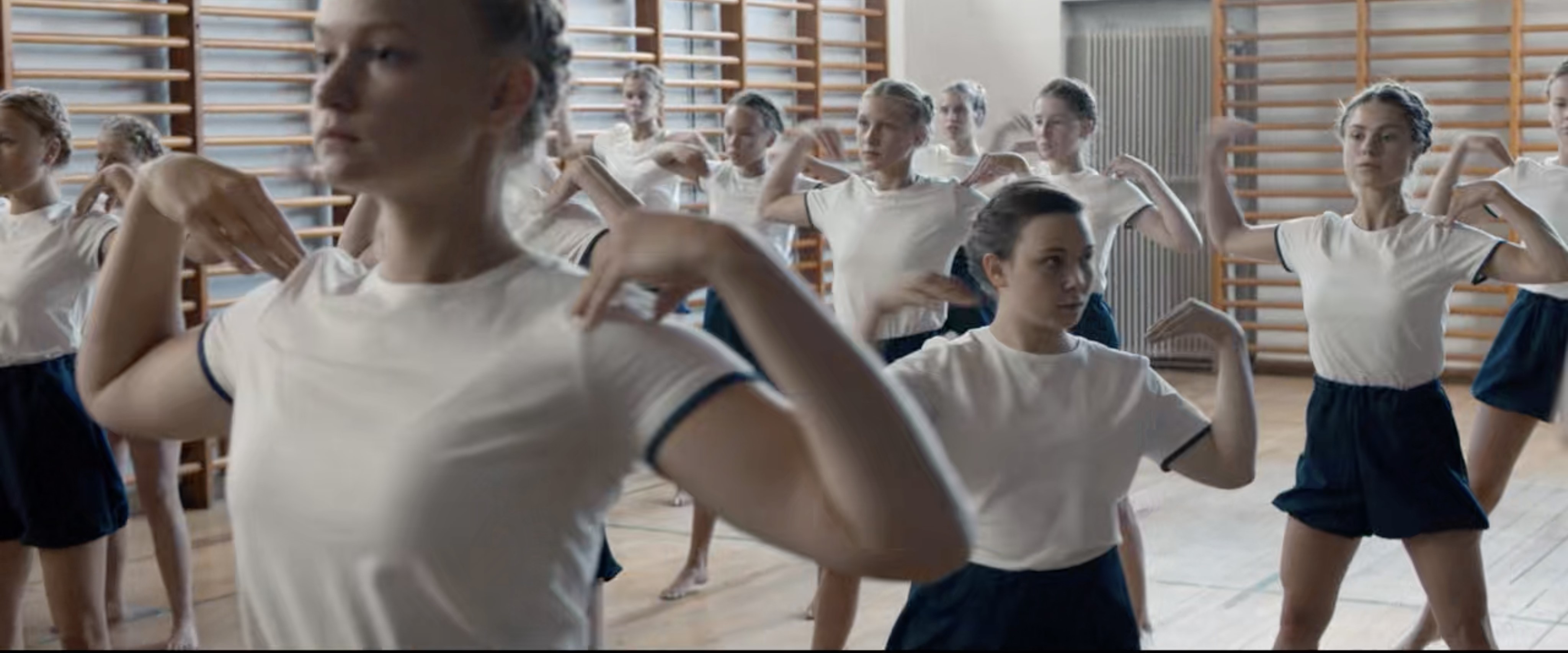 “A room of blonde-haired girls with arms bent to touch hands to shoulders face the camera. A shorter girl with darker hair is turned and looking to the right side of the frame.”