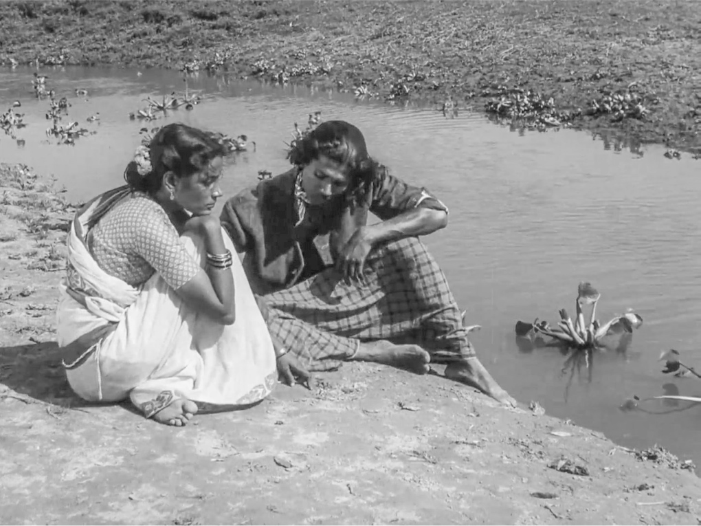 A young woman in a white sari and a young man dressed in a loincloth and shirt are pensively seated on the bank of a stream, as a few water hyacinths gently flow past them.