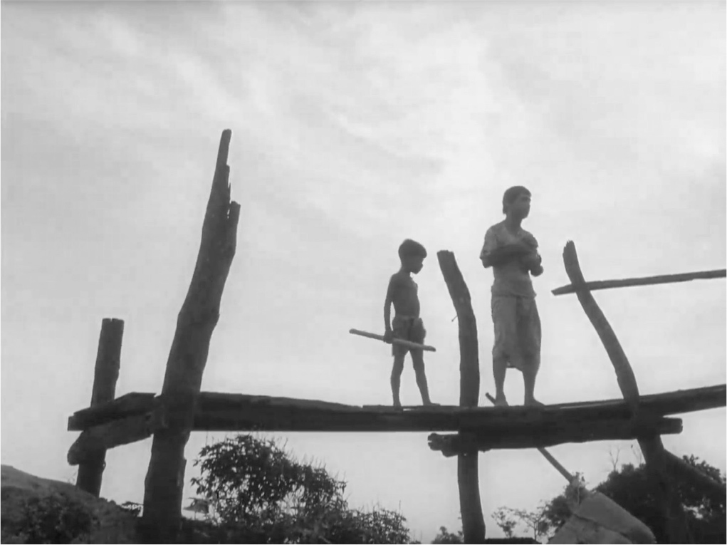 A low angle shot of a picturesque, roughly made, misshapen wooden pier shown in silhouette against the sky. A man is standing on the pier holding a baby, with a child beside him.