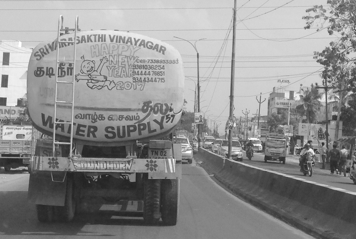 This photo shows the back of a tanker truck driving along a busy urban road. The words “water supply” and a cartoonlike image of a happy child are painted on the truck.
