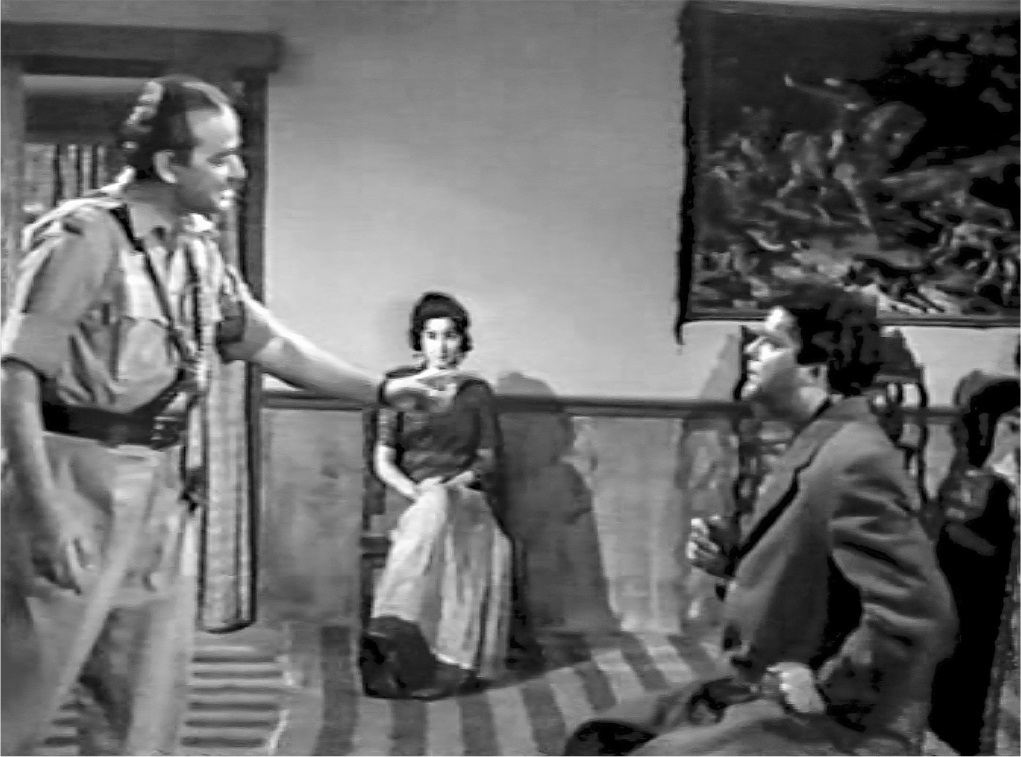 An interior scene showing a police chief in uniform standing in profile on the left, pointing towards a seated and startled man. In the center, at some distance behind them, a woman in a sari sits facing them.