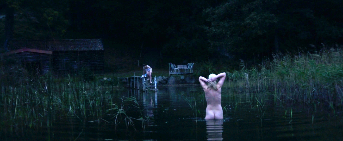A blond-haired woman stands nude in a lake and watches as a lightly clad, dark-haired woman steps into the water as well.