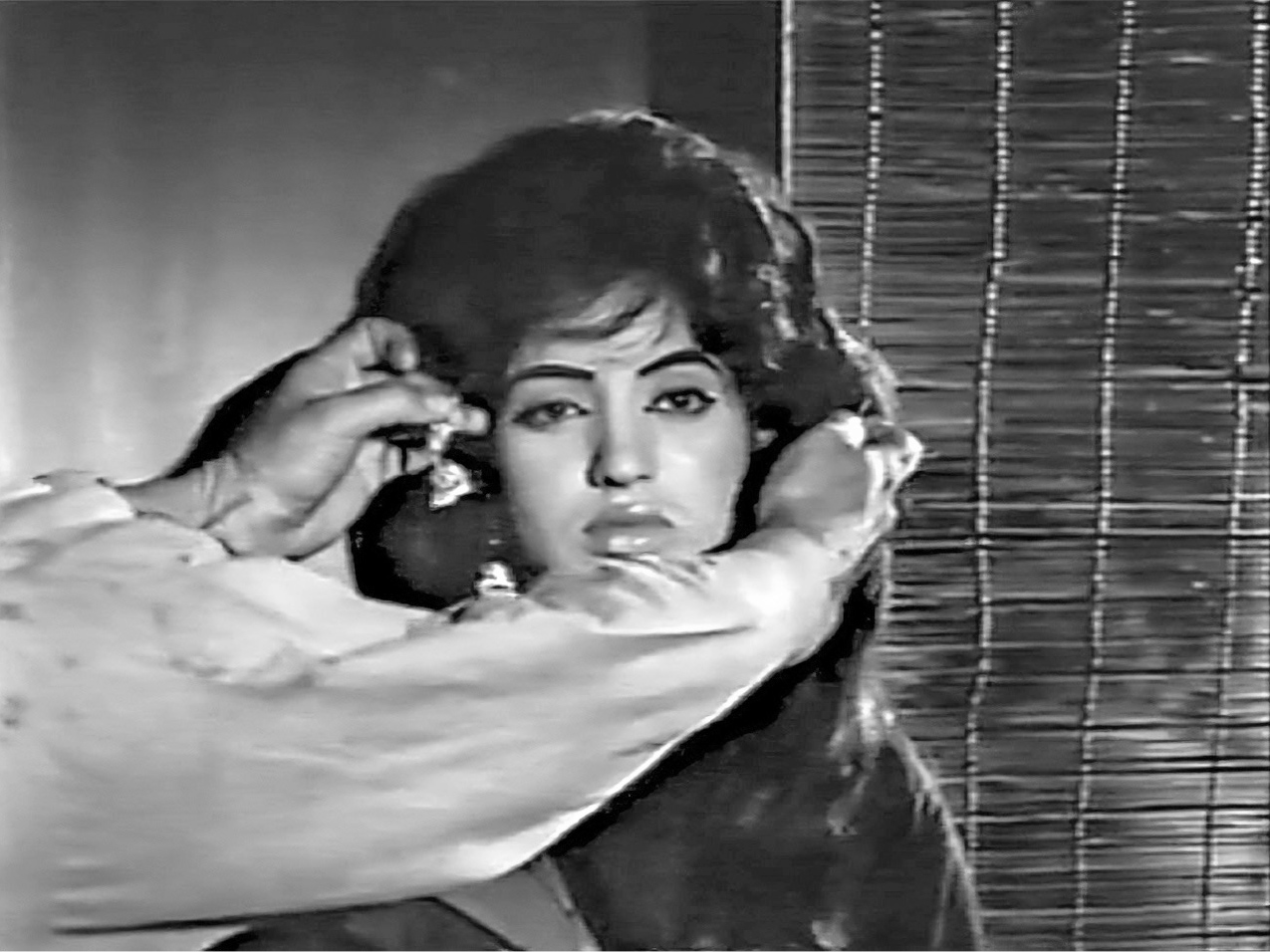 An interior close-up of a woman’s face, with a blank expression, wearing pendant earrings. The two arms of a man standing outside the frame stretch towards her face from the left, holding another set of pendant earrings.