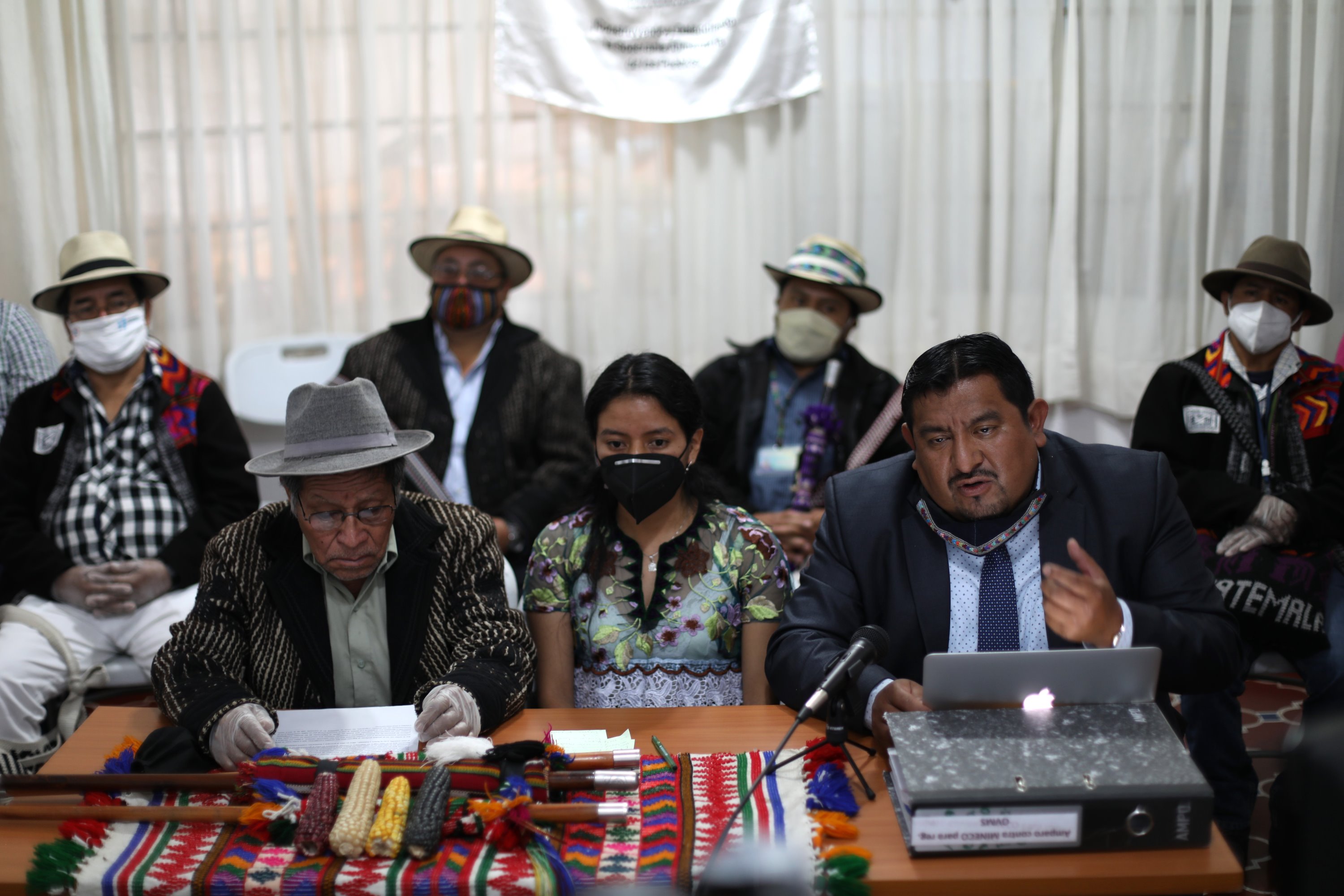 In curtained room, six Maya plaintiffs wearing pandemic masks sit behind a table with maize cobs perched on three carved wooden staffs. A man in a suit and tie seated behind a laptop makes a charismatic hand gesture as he speaks into a microphone.