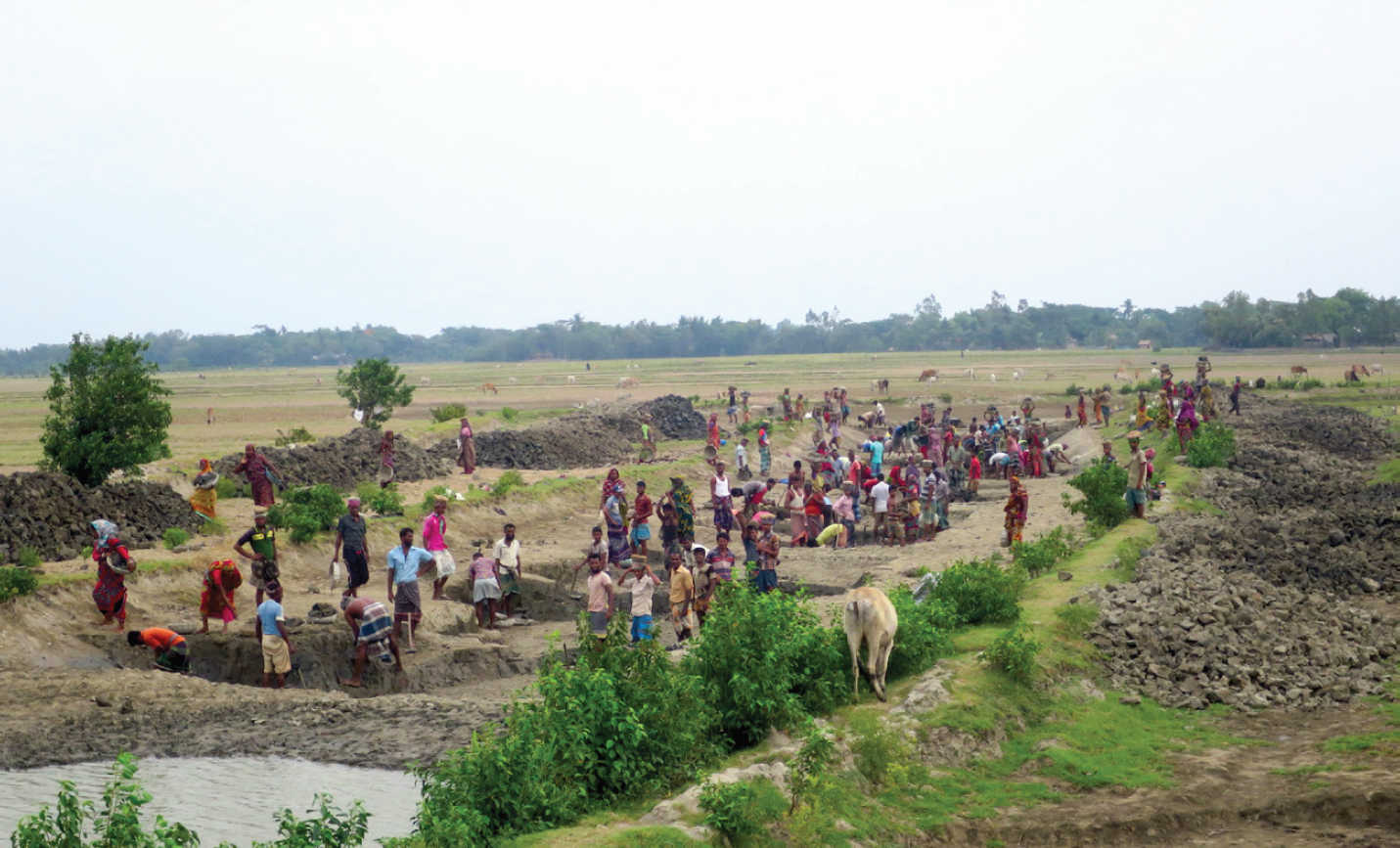 A photograph of several dozen people gathering in and around a dry canal that crosses a flat field. Some are digging, and there are mounds of dirt on either side.