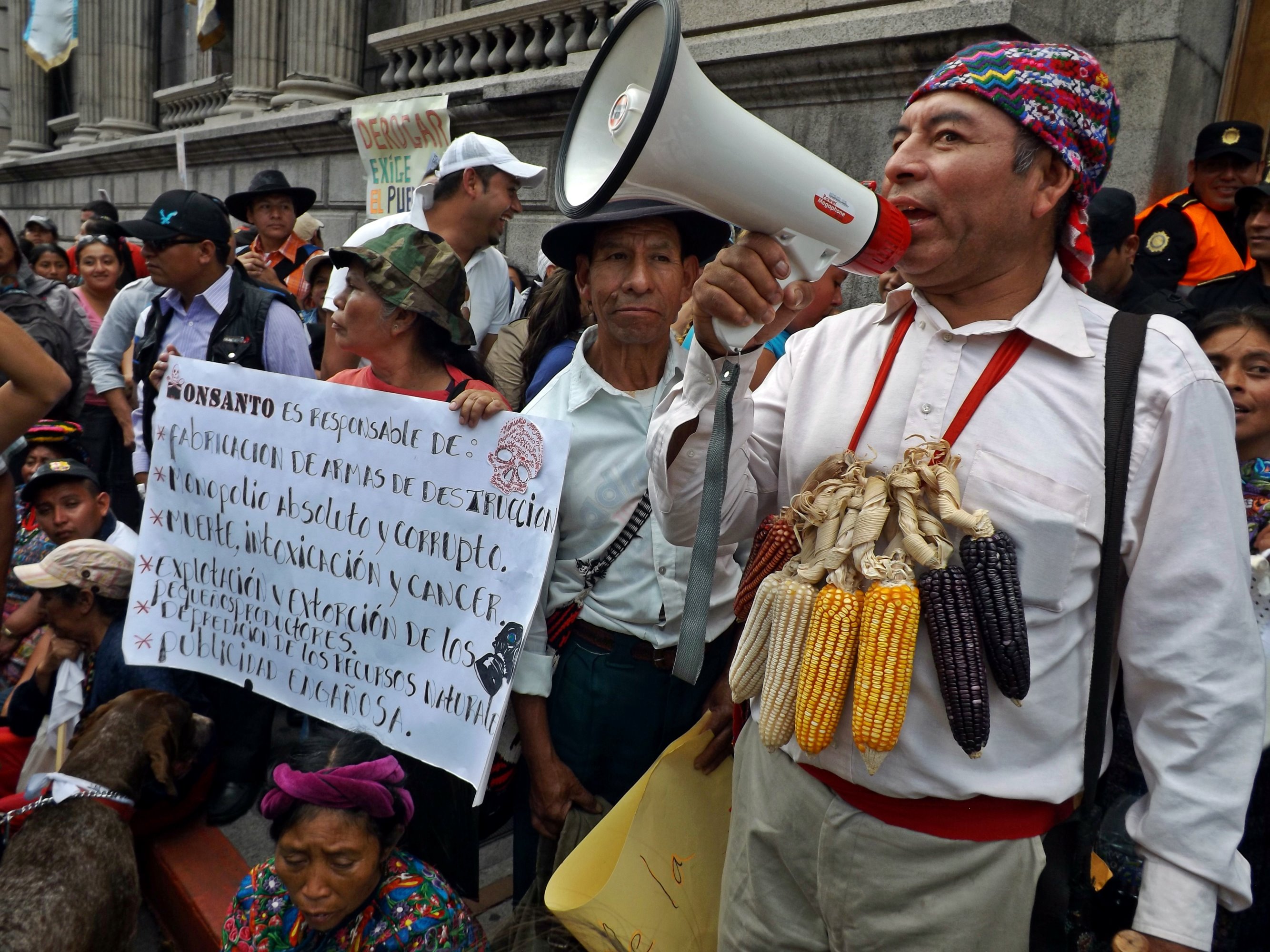 Maya man with a head scarf wearing a “necklace” of maize cobs speaks into a bullhorn in a crowd of Maya protestors carrying banners outside a colonial-style building.