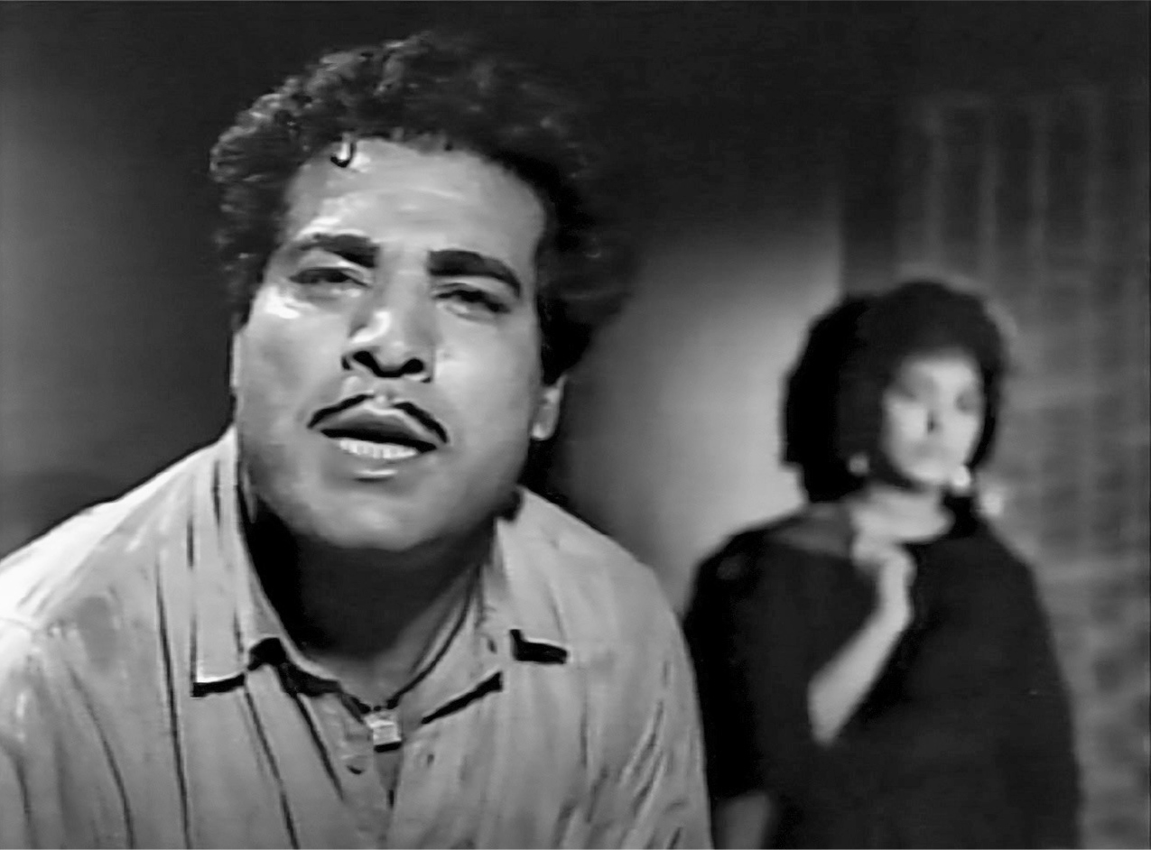 An interior close-up of the impassioned face of a man facing the camera, with a woman in the background on the right.