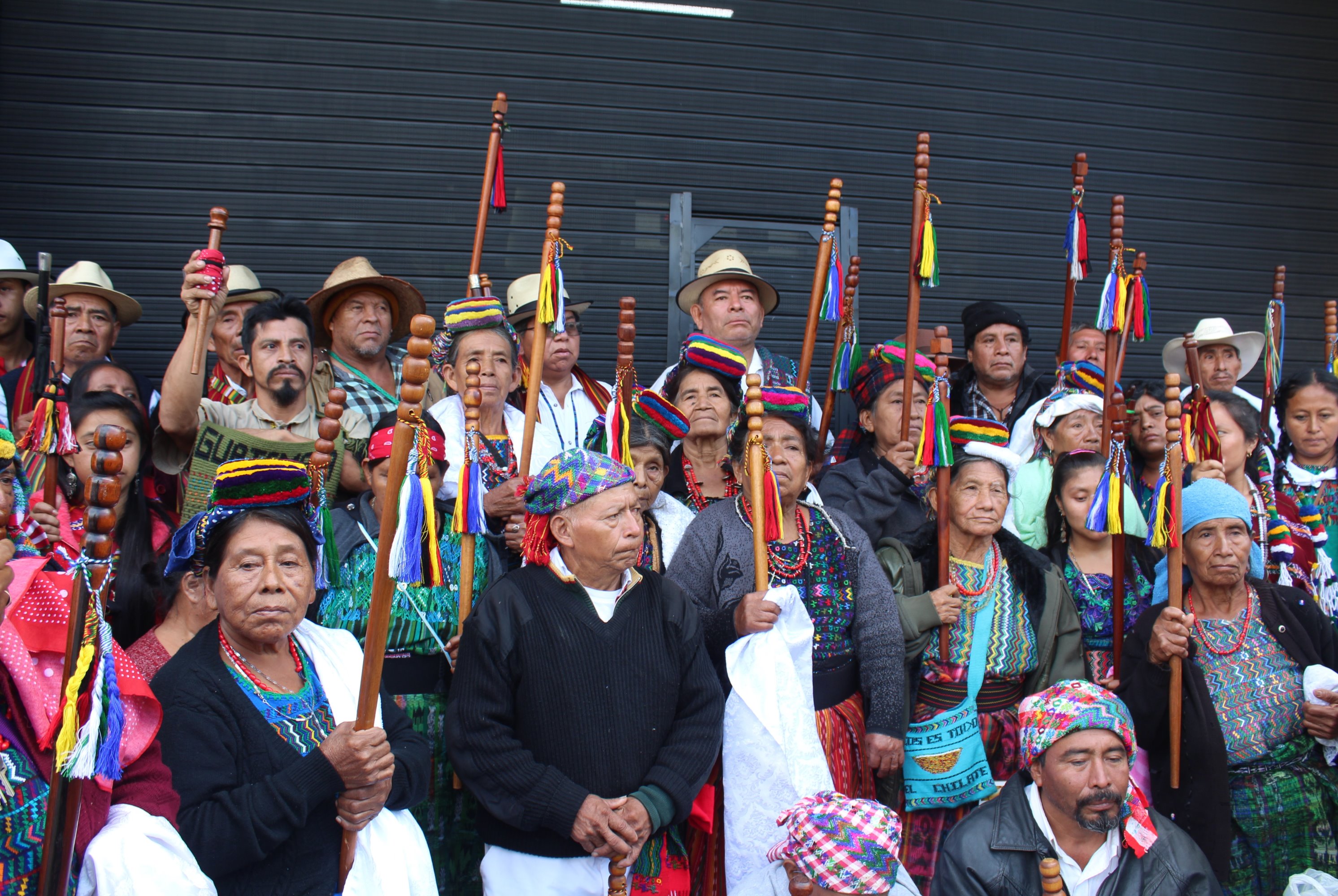 Male and female elders hold carved wooden staffs with tassels and pose for a photo outside of a city building.
