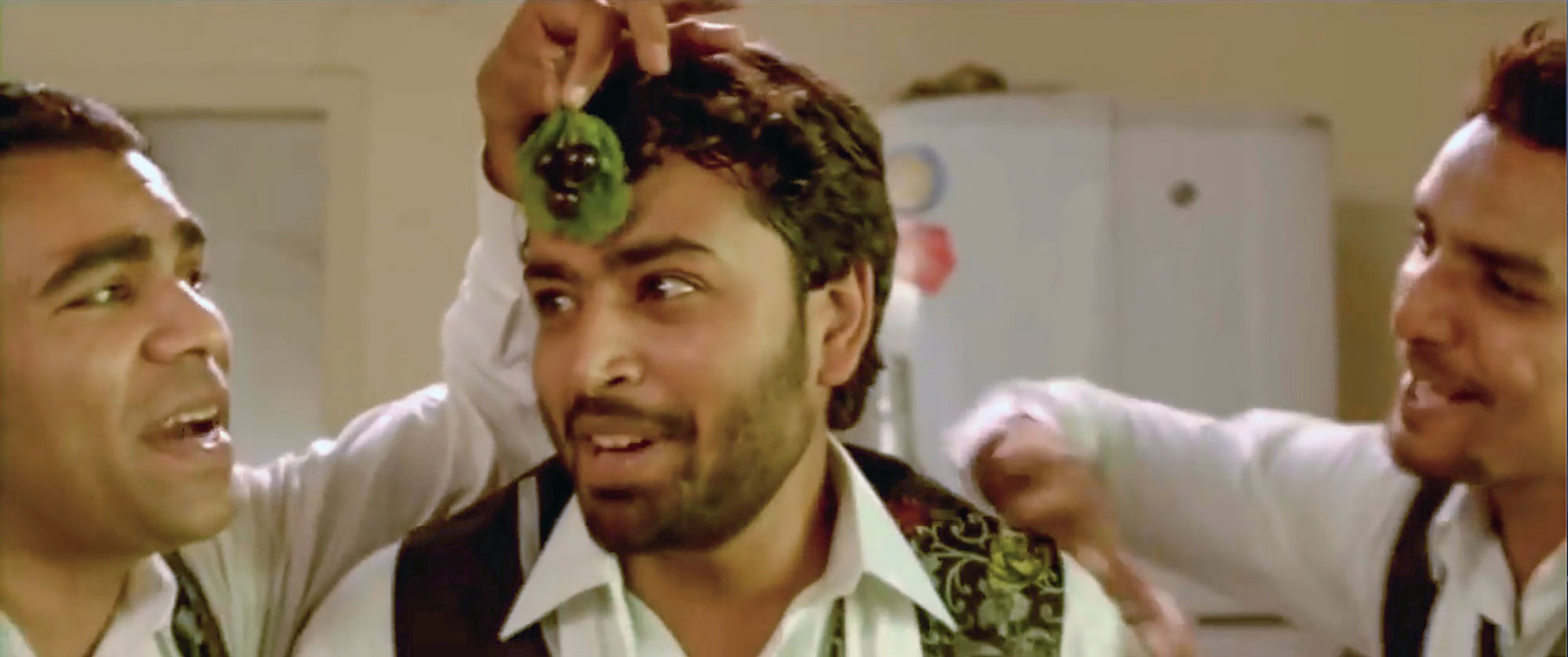 A close-up interior shot shows the faces of three young men dressed as waiters and dancing. The man on the left holds a leaf and small bunch of grapes reminiscent of a jeweled pendant on the forehead of the central figure.