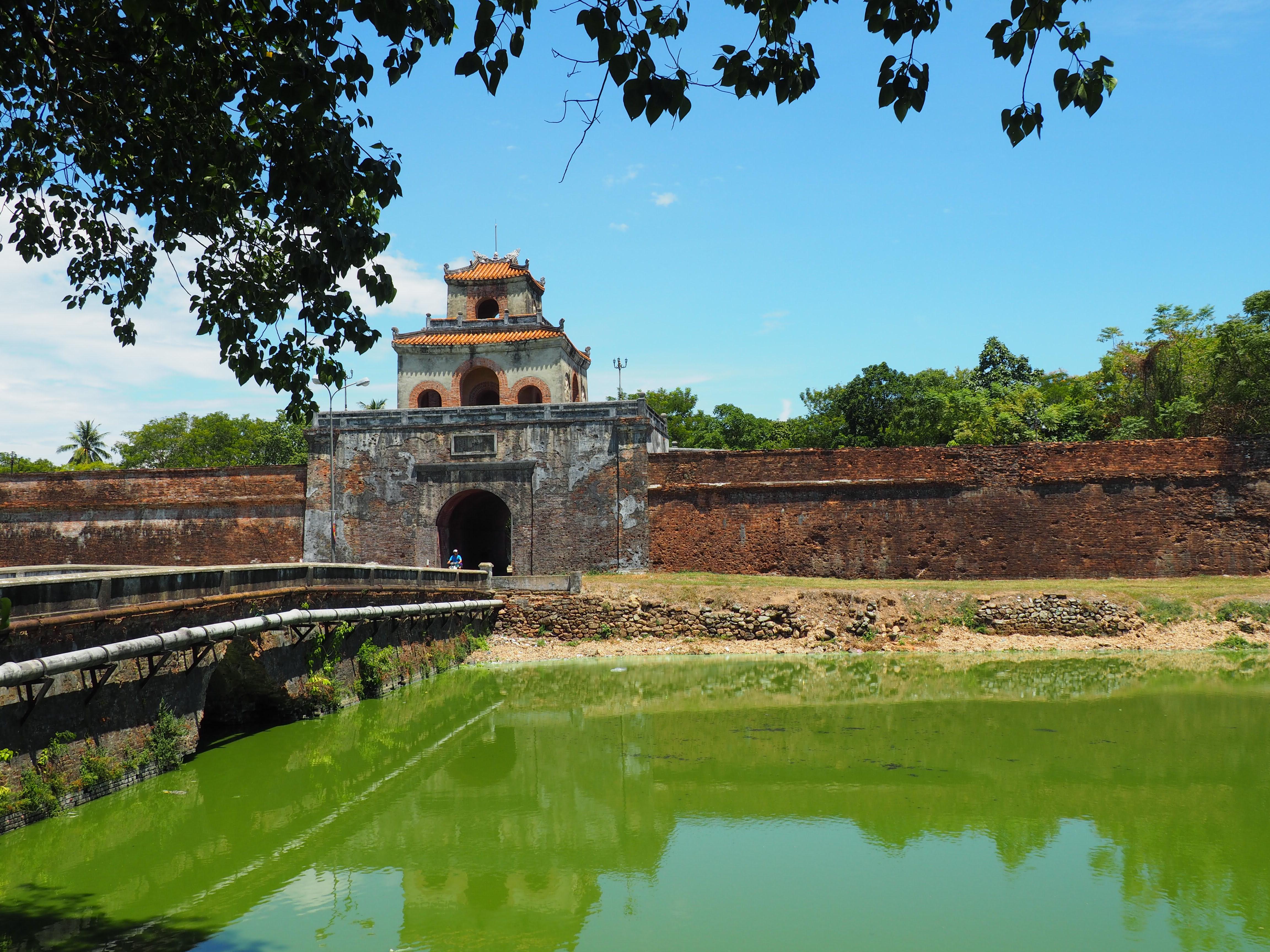 A long stone wall flanking a large, three-tiered, pavilion-style gate, viewed from across a channel of water.