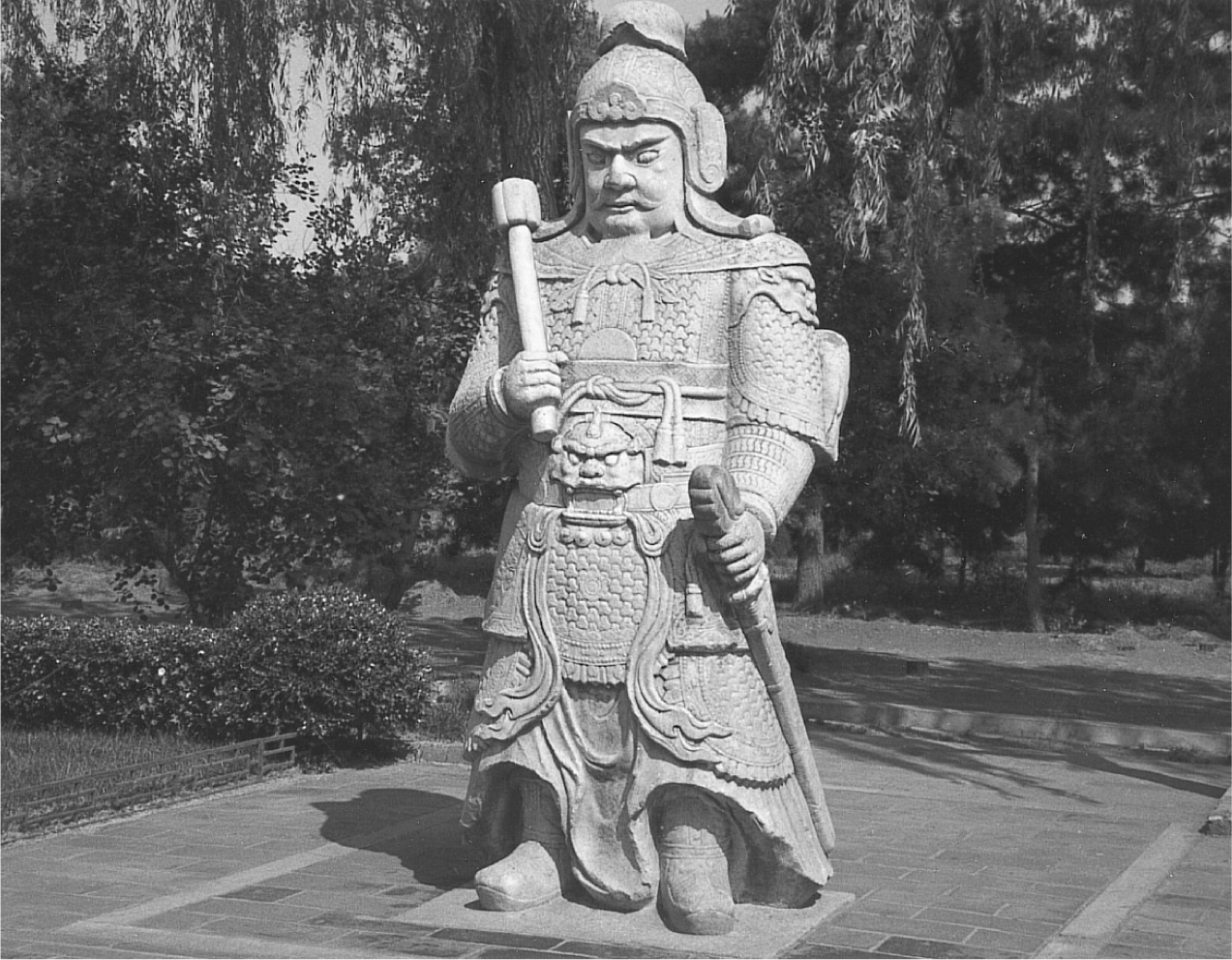 A photograph of a stone statue of a sturdy, stern warrior, armored and helmeted, with a sword in one hand and a club in the other, and a glare in his eyes.