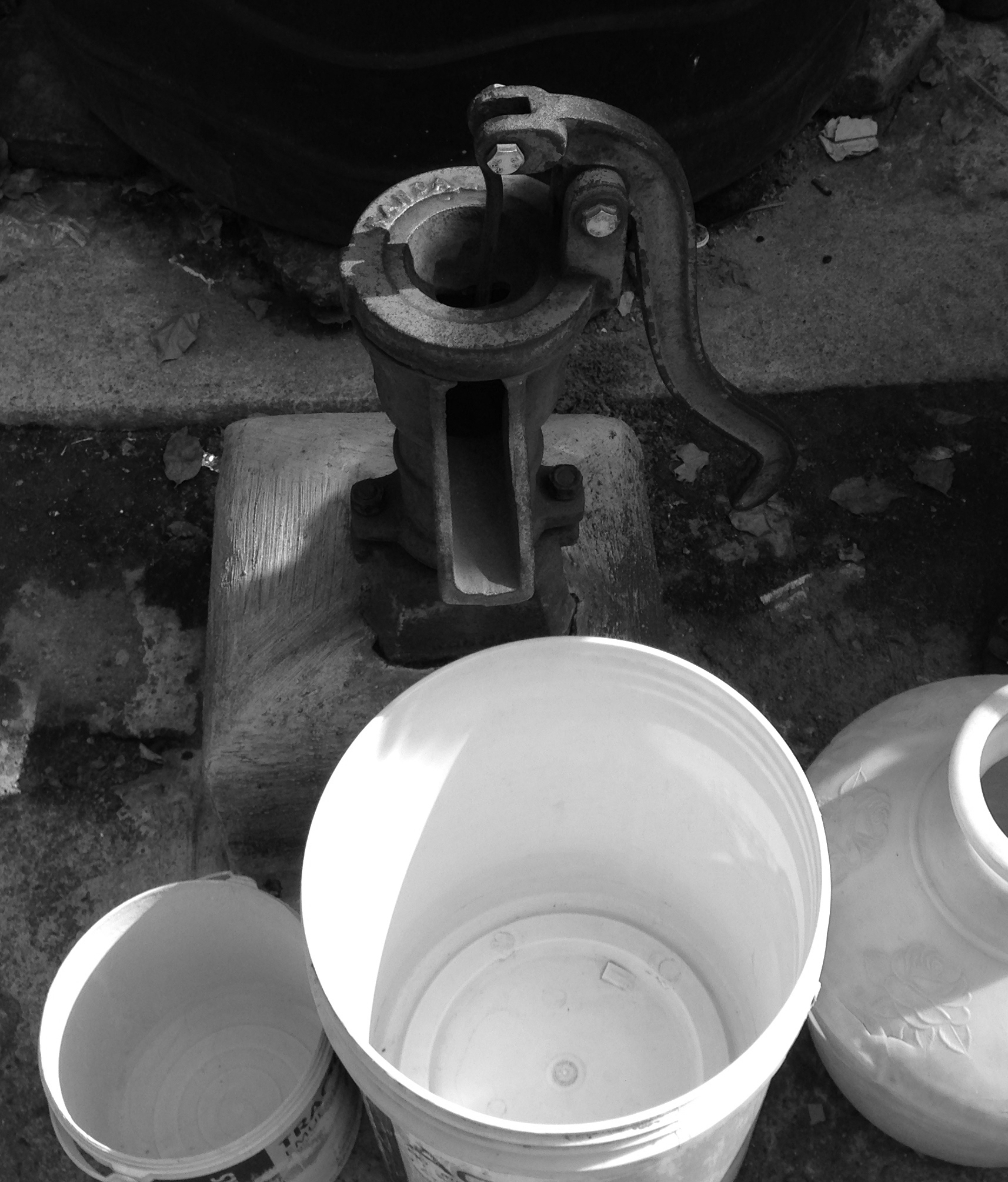 This photo shows three empty white buckets set on the ground beside a metal water pump.