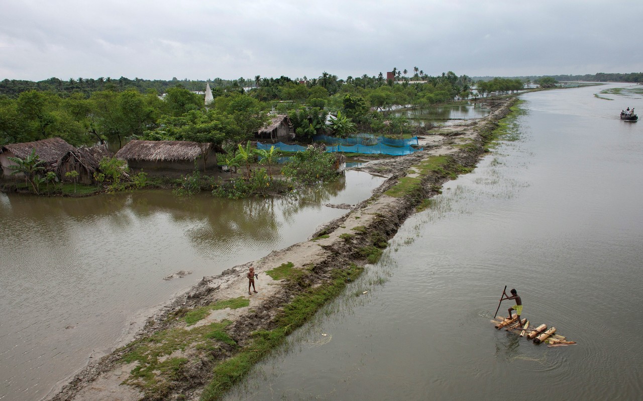 A photograph of a swollen river bordered by a narrow embankment of land that separates it from a flooded field. A man on a small raft is poling it towards the embankment where a child waits.