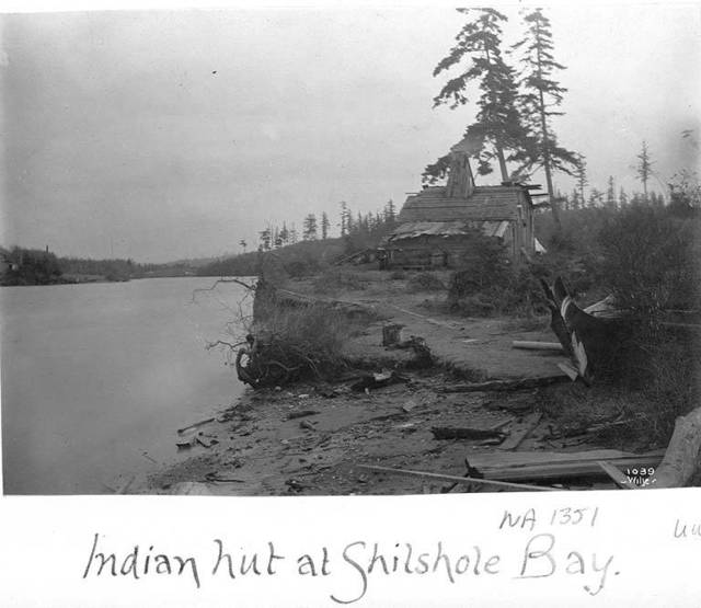 View of bay and beach, in foreground is painted prow of canoe, in background is small wooden house