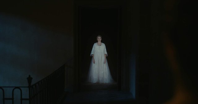 A screenshot from "Portrait of a Lady on Fire" depicting a woman in a white dress (Haenal) standing at the end of a dark hallway, with the bottom of the dress fading into the darkness around the figure.