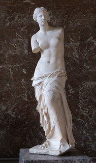 The Venus de Milo is an ancient Greek statue of Aphrodite, the goddess of love and beauty. The sculpture stands over 6 feet tall and is crafted from marble. It depicts a partially draped female figure with her upper body exposed and her lower body covered in flowing drapery. The sculpture is missing both arms and features detailed, wavy hair pulled back and a serene facial expression. The body’s contours are finely carved.